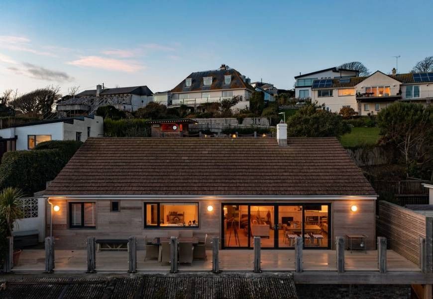 Coastal home with deck and sliding glass doors, illuminated at dusk. Other homes on a hillside.
