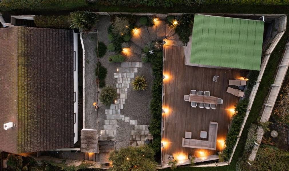 Overhead view of a backyard with a stone patio, wooden deck, and a green-roofed structure, lit by string lights.