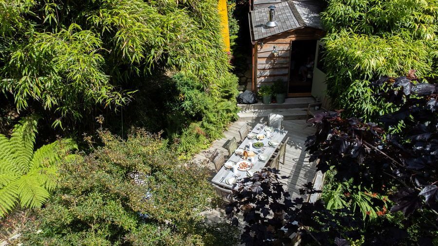 A garden patio with a table set for a meal, surrounded by lush greenery, leading to a small wooden structure.