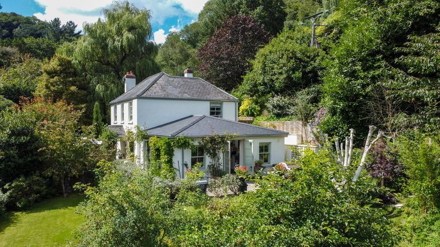 White house nestled in lush, green hillside. Trees and vegetation surround the two-story building.
