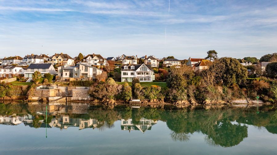 Row of waterfront houses, trees, and water with reflections under a blue sky.