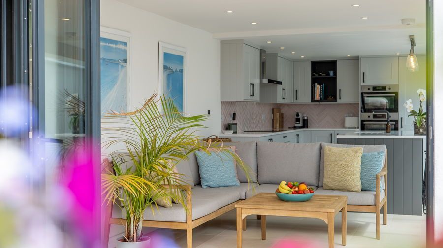 Lounge area opens to kitchen. Light wood sofa with blue and yellow pillows, small table with fruit.