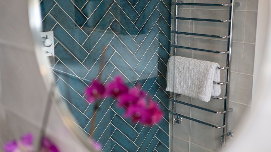 Oval mirror reflects a bathroom with a teal herringbone tile wall, a towel warmer with a white towel, and purple orchids.