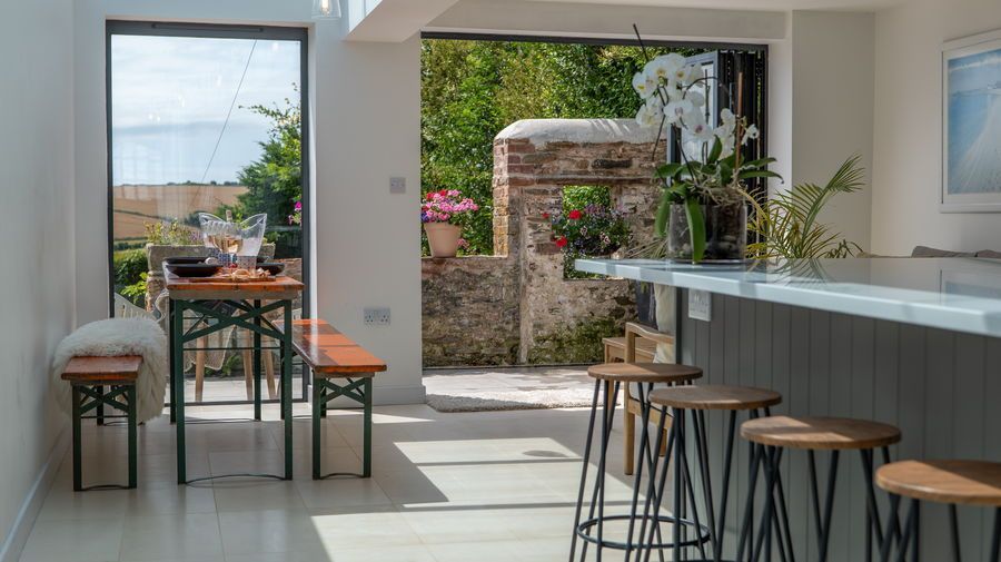 Kitchen with bar stools, table, and view to a garden through open door. Sunlight streams through.