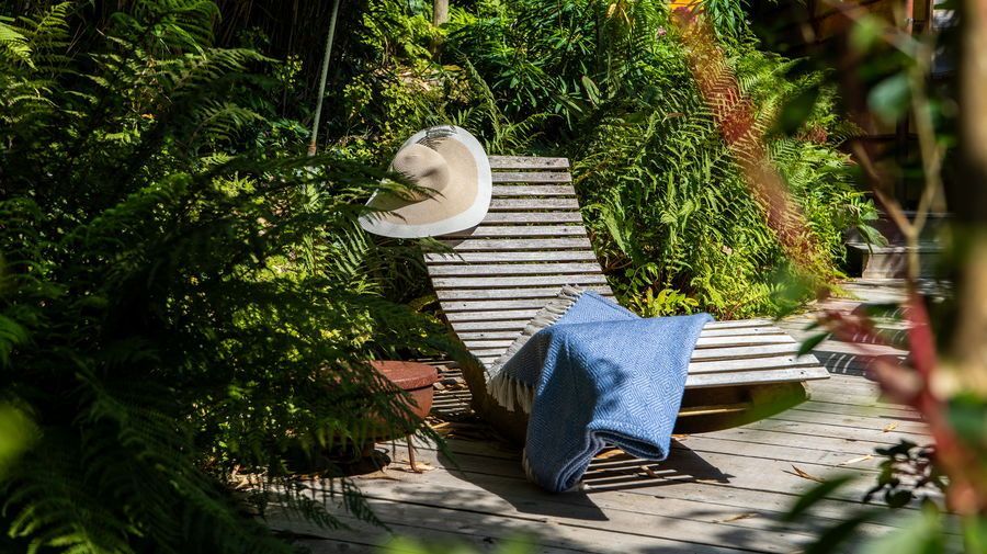Wooden chaise lounge with hat and blue blanket in a lush green garden setting.