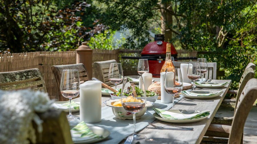 Outdoor dining table set for a meal, featuring plates, glasses, and a red grill in the background.