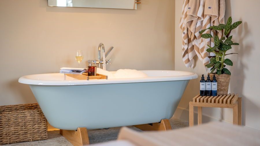 Blue bathtub with bubbles, tray with drinks, and toiletries in a light, neutral bathroom.
