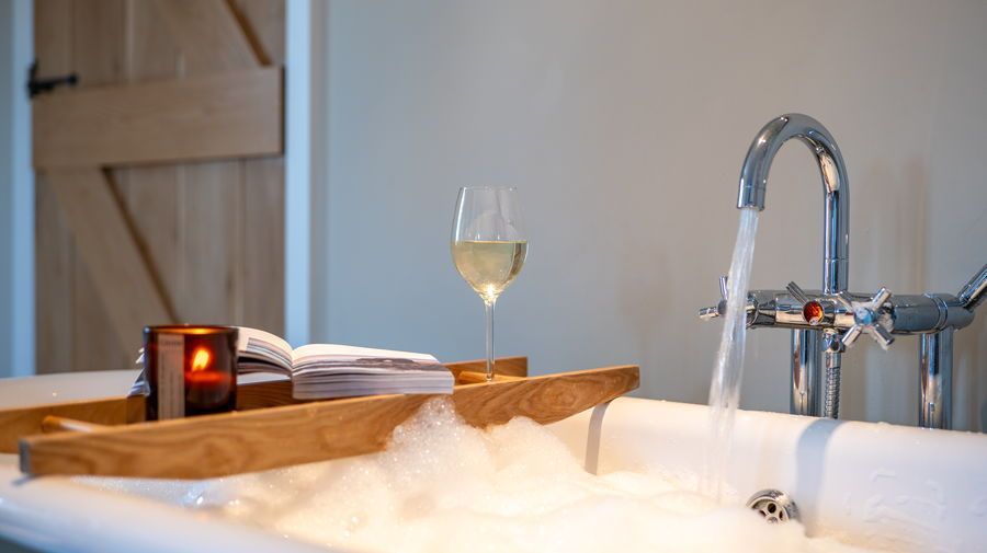 Bathtub with bubbles, a wooden tray holding a glass of wine, a candle, and towels. Water flows from a chrome faucet.