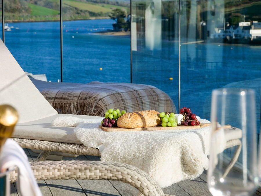 Lounge chair with food and drink overlooking a bay. Glass windows, water, and greenery in the background.