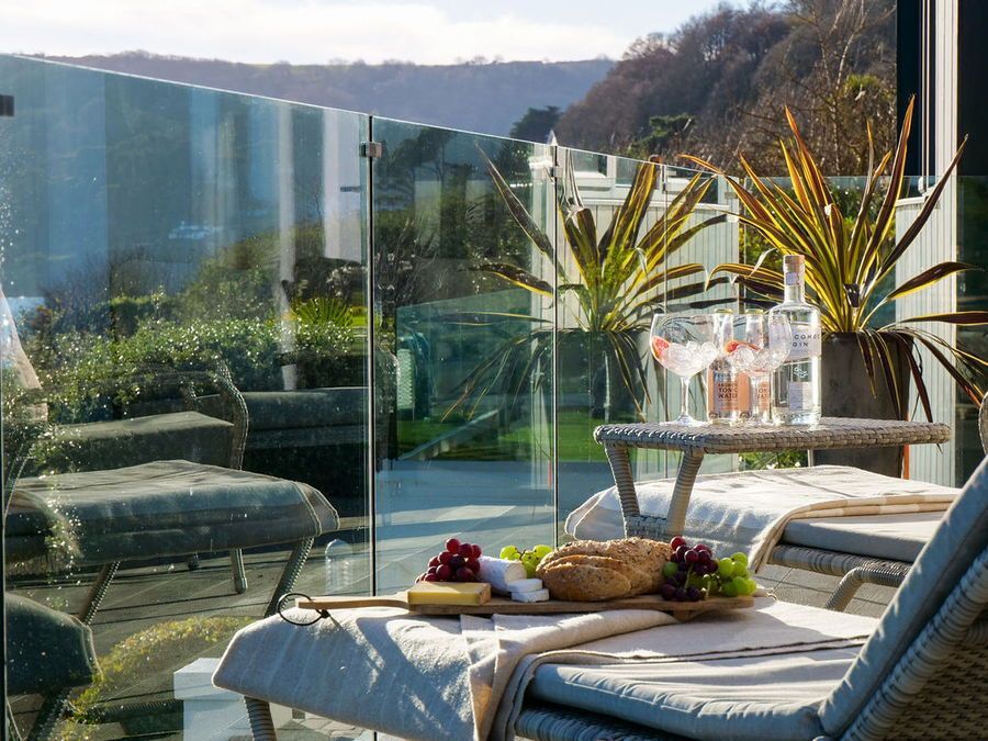 Patio scene: cheese board and drinks on small table, lounger, glass railing, hillside view.