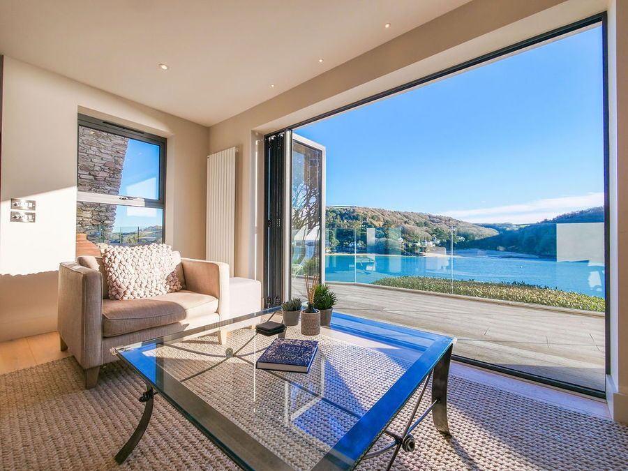 Living room with water view, beige walls, glass table, beige sofa, folding doors open to blue water, sunny day.