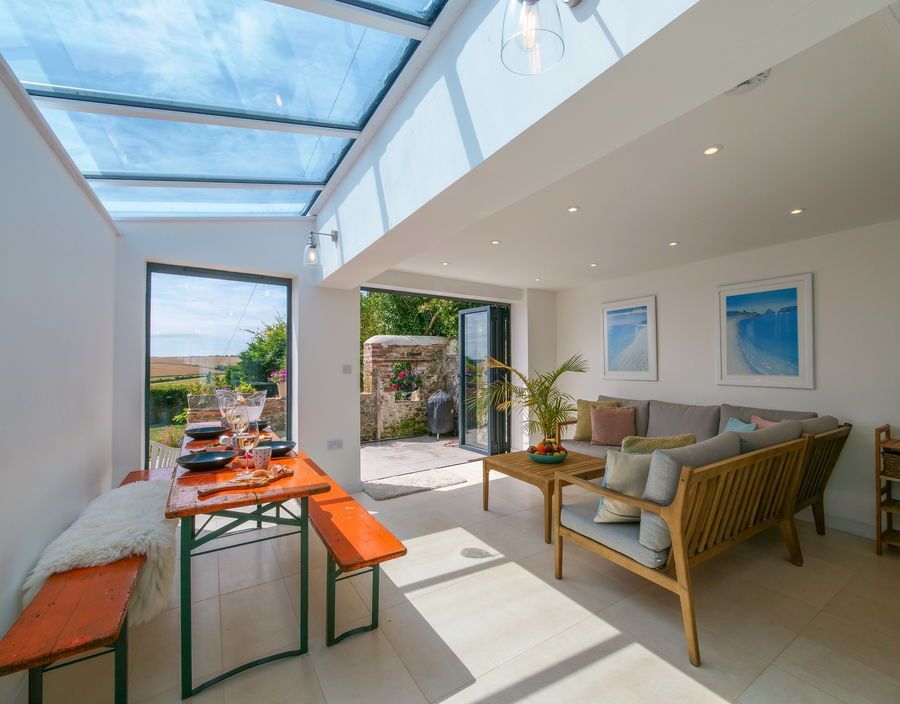 Bright sunroom with glass roof and open doors. Dining table, wicker furniture, and artwork.