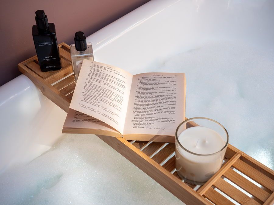Bathtub tray with book, candle, and toiletries in a bubble bath.