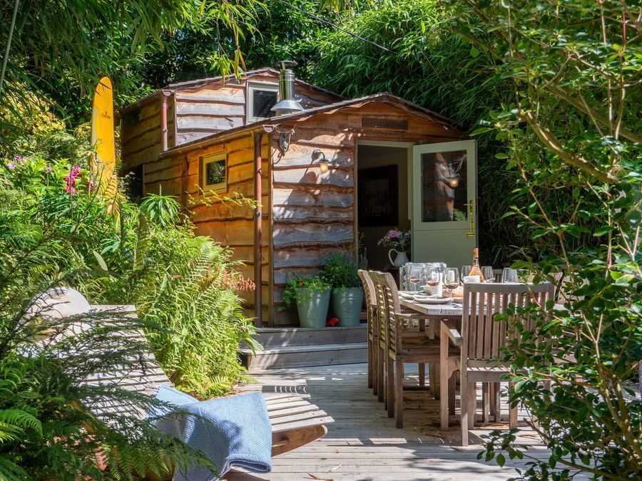 Wooden shed with open door, outdoor dining table, and lush greenery in a sunny garden setting.