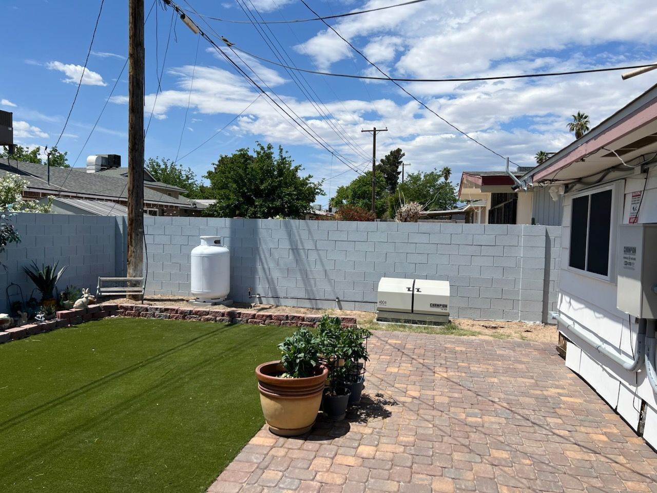 Backyard with artificial turf, brick patio, and a propane tank against a gray block wall under a blue sky.