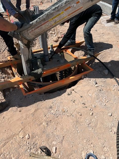Concrete being poured into a hole by workers. Orange construction equipment and dirt are visible.