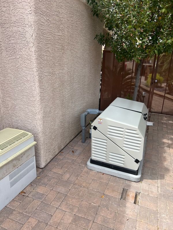 Tan-colored house exterior with an electrical generator on a brick patio. A tree and brown fence are visible.