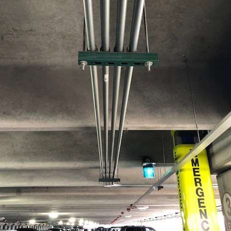 Pipes hanging from a concrete ceiling in a parking garage. Emergency signage and blue light are also visible.