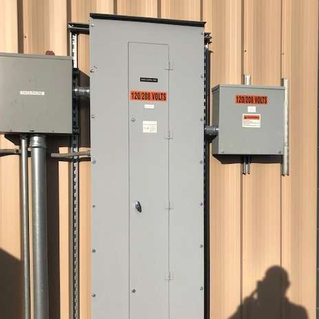 Gray electrical panel with warning labels and connected boxes on a beige building exterior.