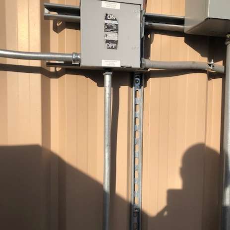 Electrical panel box mounted on a beige metal wall with conduit pipes and metal support.