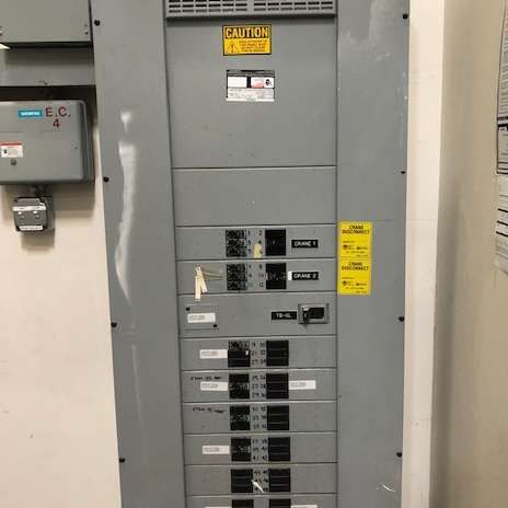 Gray electrical panel with several circuit breaker rows in a building. Warning labels are visible.