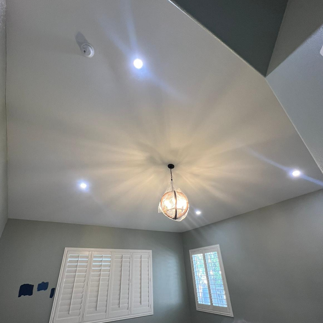 Ceiling with recessed lights and a hanging chandelier, room with window and shutters, gray walls.