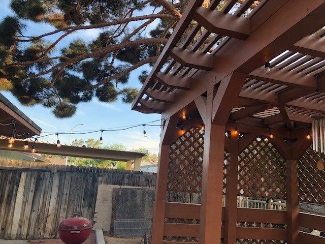 Brown wooden pergola with string lights, backyard setting, blue sky, and a grill.