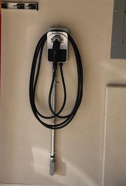 EV charger mounted on a beige wall, black cable looped, metal arm, gray electrical panel nearby.
