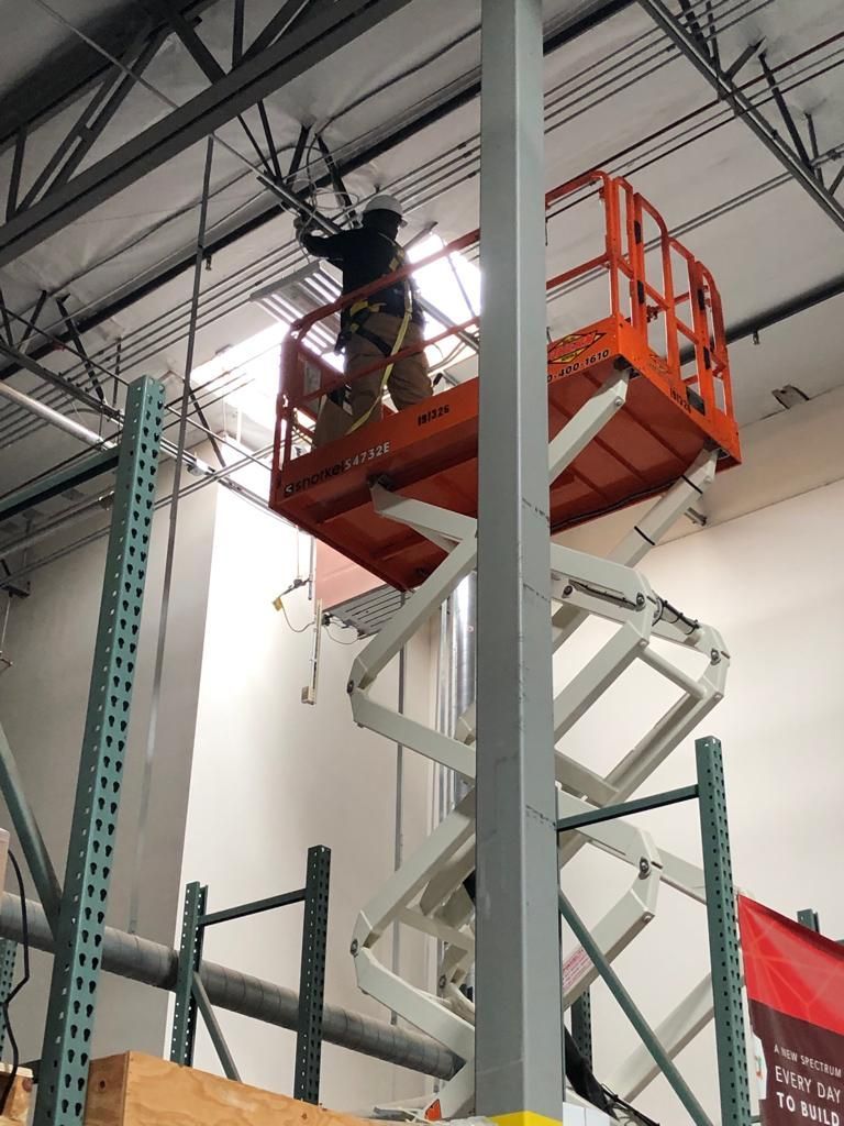 Person on orange lift, working on overhead wires in a warehouse, with visible shelving and support beam.