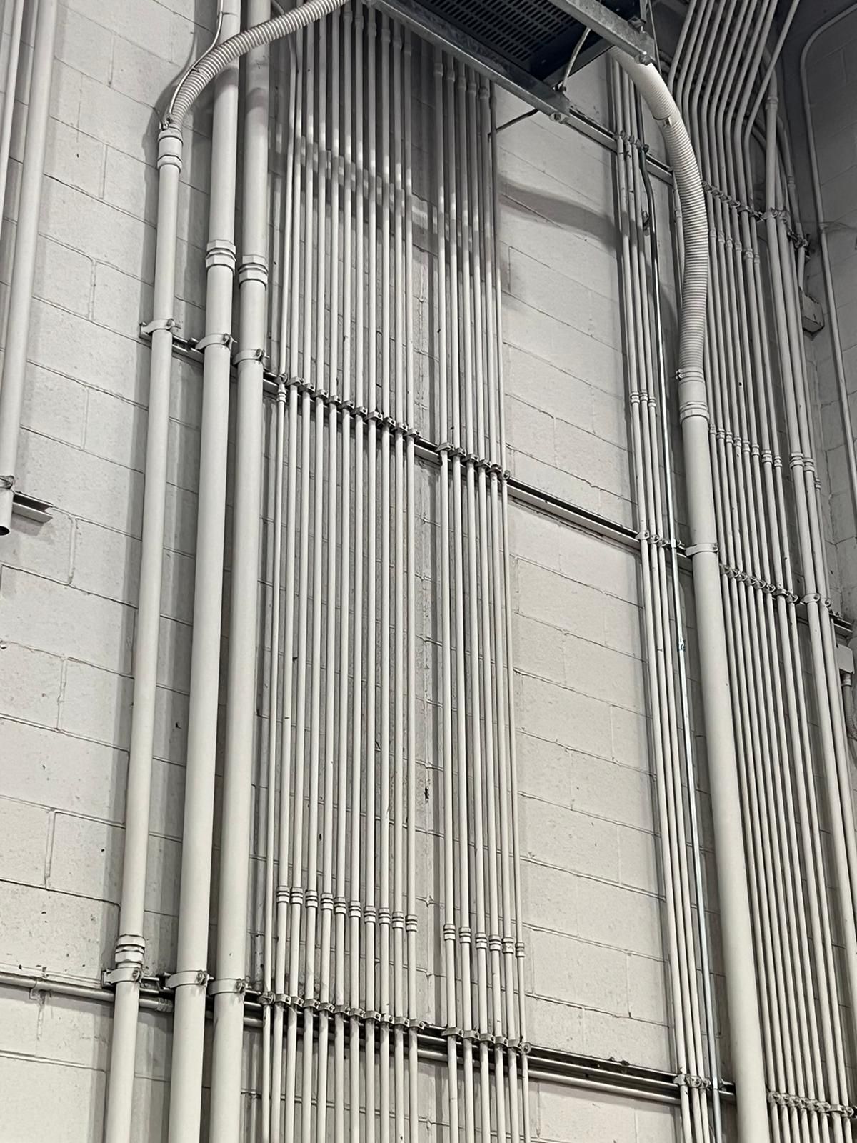 Metal conduits and supports run vertically and horizontally on a concrete wall.
