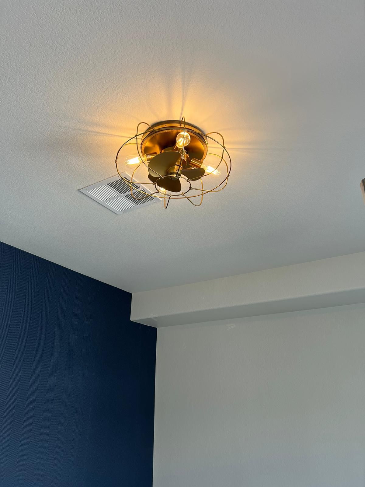 Ceiling fan with gold finish, lit, mounted on a white ceiling. Blue and white walls in the room.