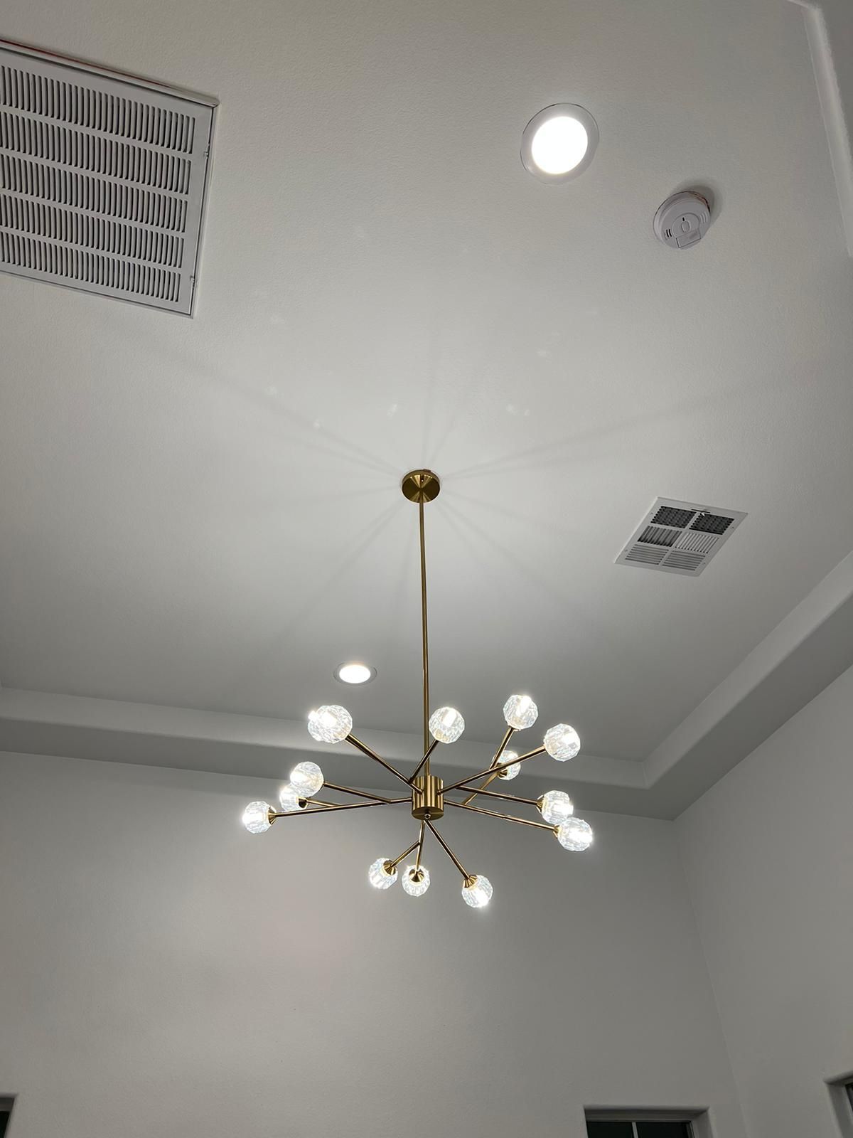 Gold chandelier with spherical lights hanging from a white ceiling, alongside a vent and smoke detector.