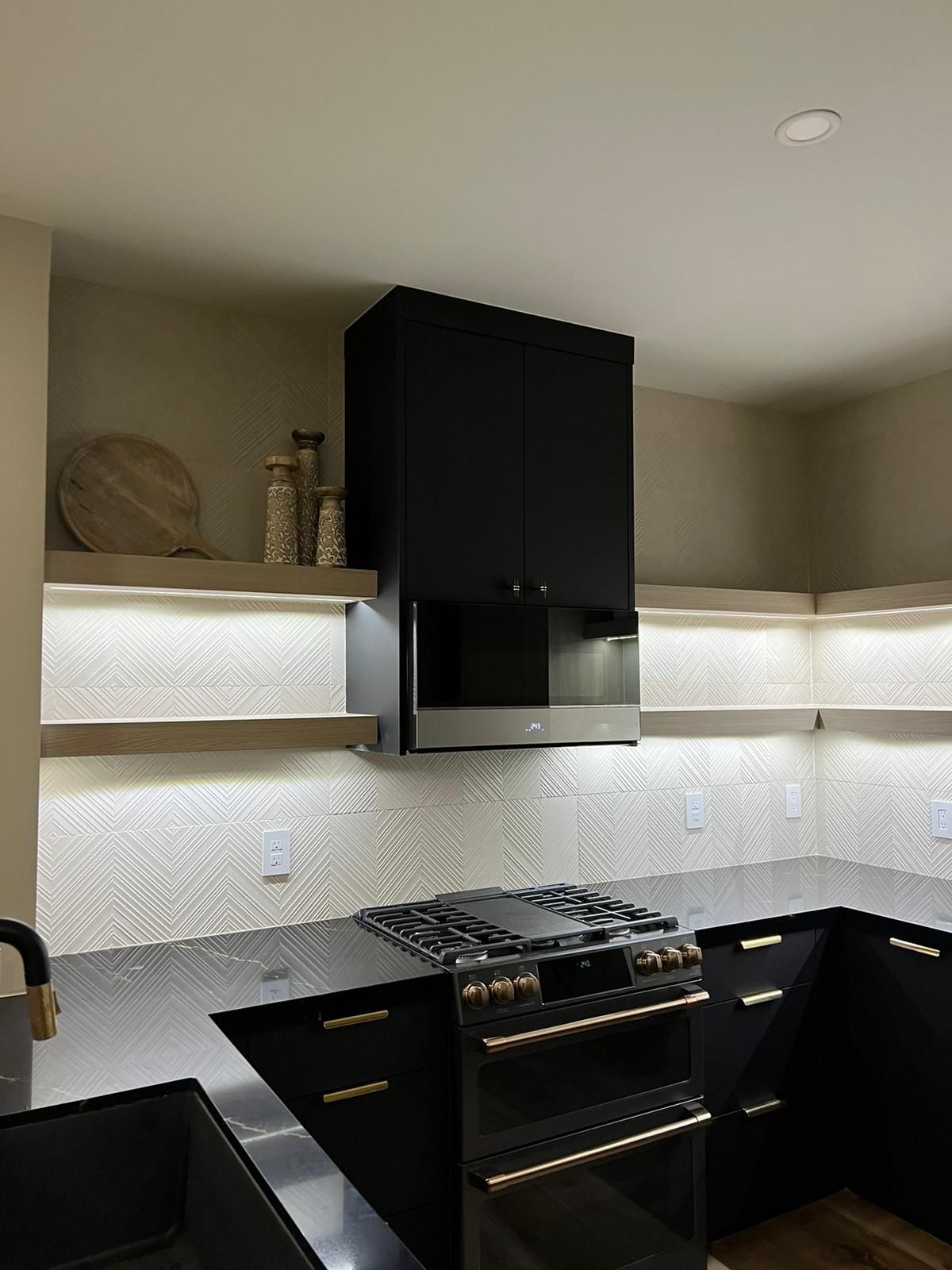Black kitchen with stove, cabinets, and decorative shelves. Backsplash with a geometric design.