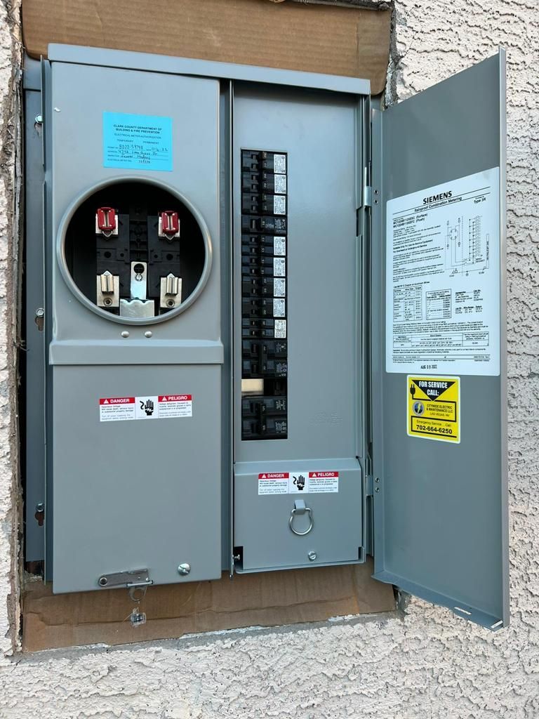 Gray electrical panel with open door, breakers, meter socket, and warning labels on a stucco wall.