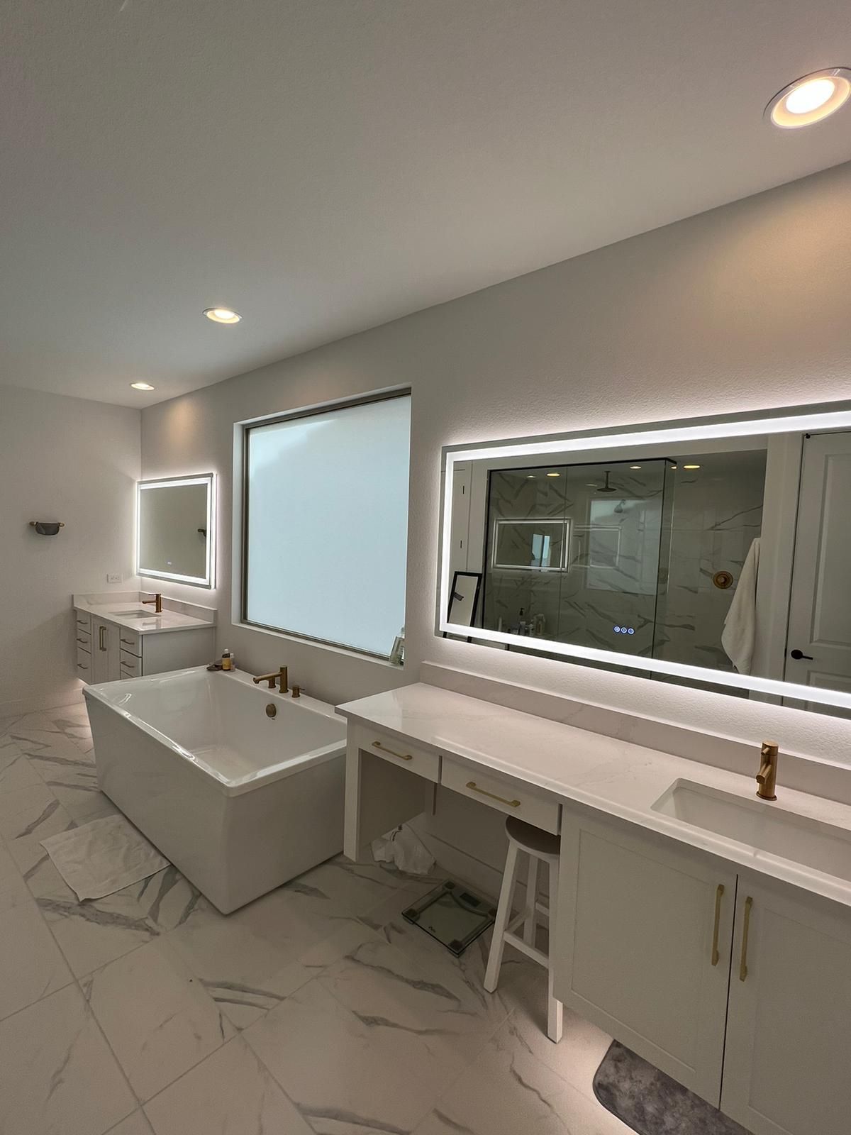Modern white bathroom with large mirrors, a tub, vanity, and a window with a closed blind.