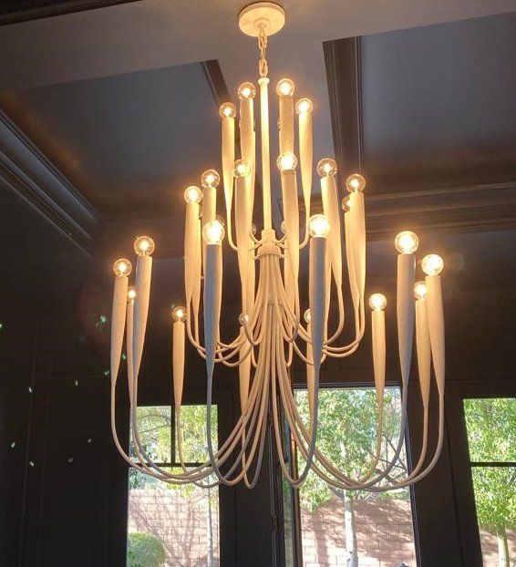 White candle-shaped chandelier with many glowing lights, hanging in a room with a dark ceiling and a window.