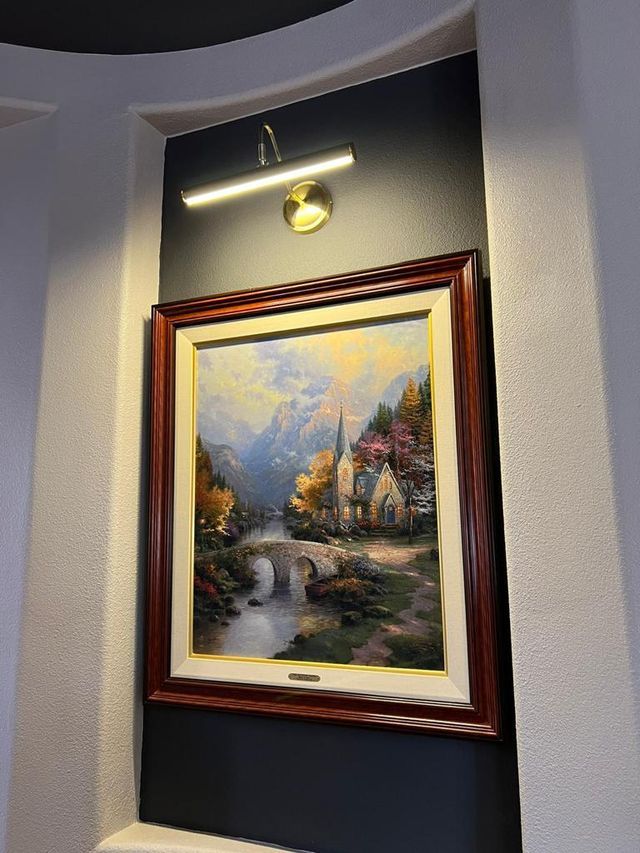 Painting of a landscape with a bridge, lit by a wall-mounted light. Wooden frame, dark wall.