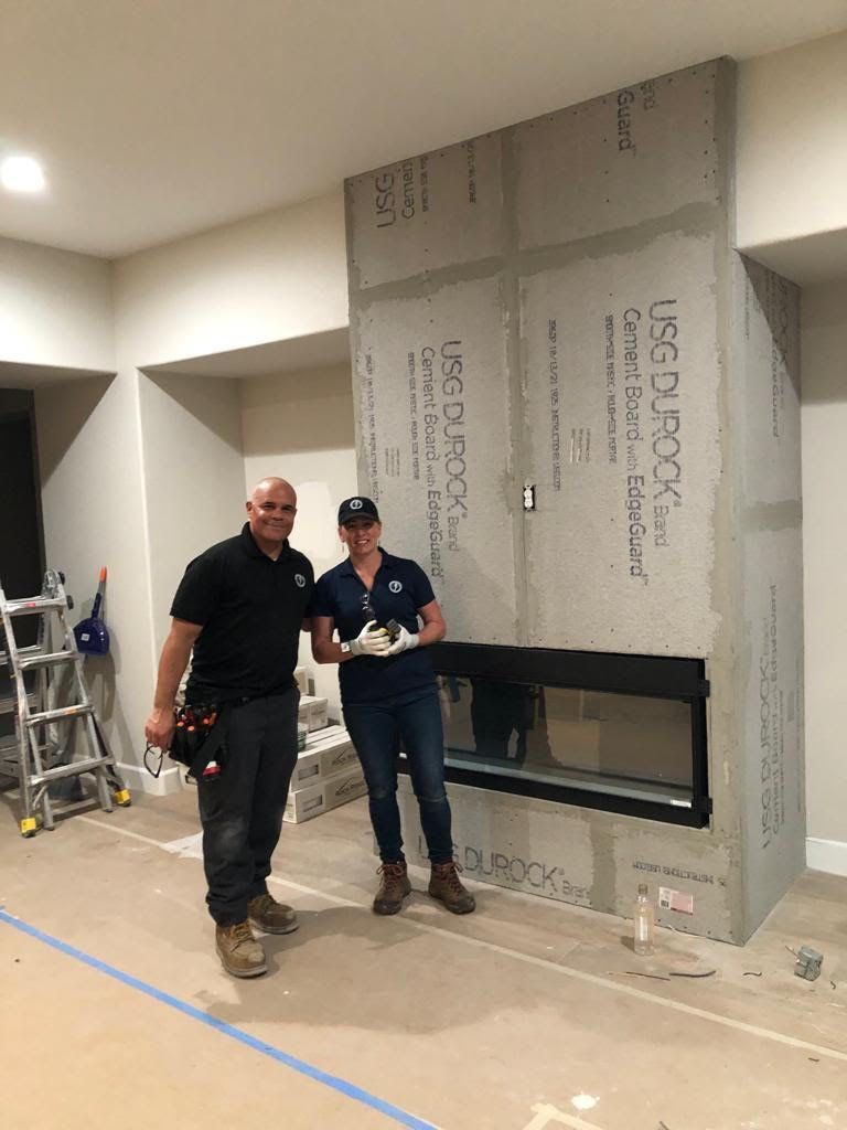 Two people stand in front of a modern fireplace. The unfinished structure is grey, and they hold tools.