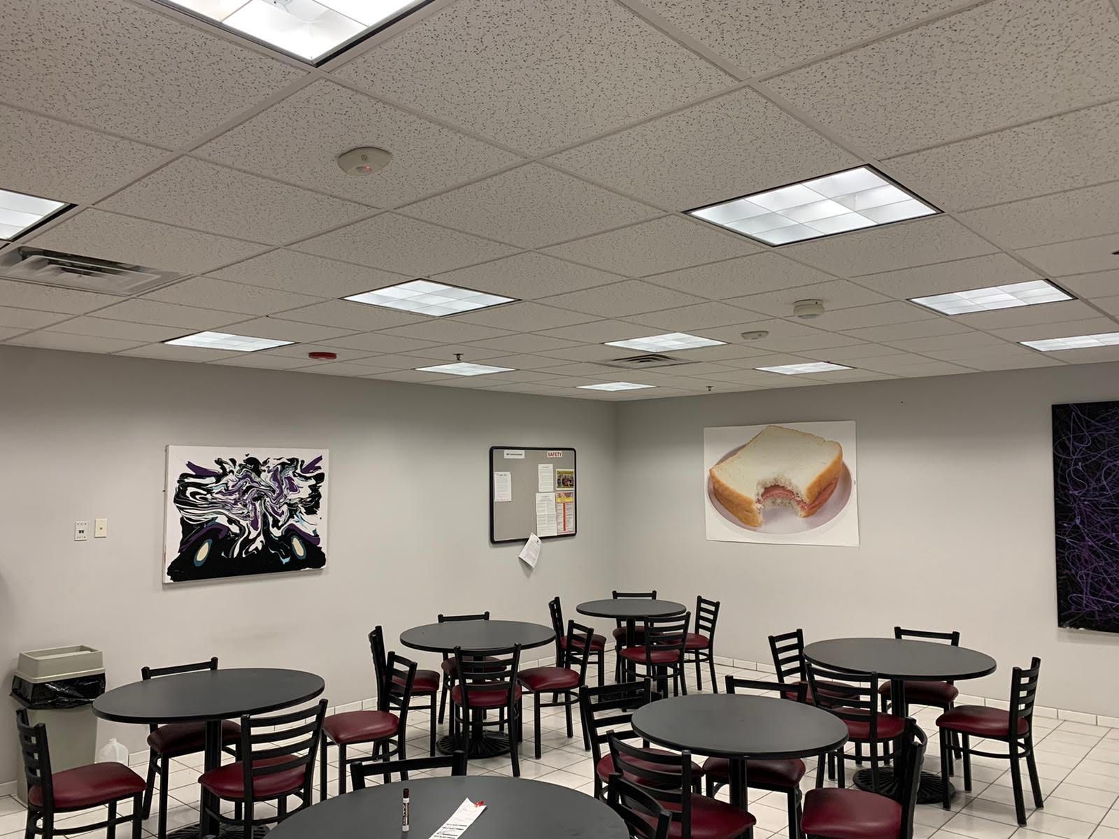 Cafeteria interior with round tables and chairs, artwork on the walls, and square ceiling lights.