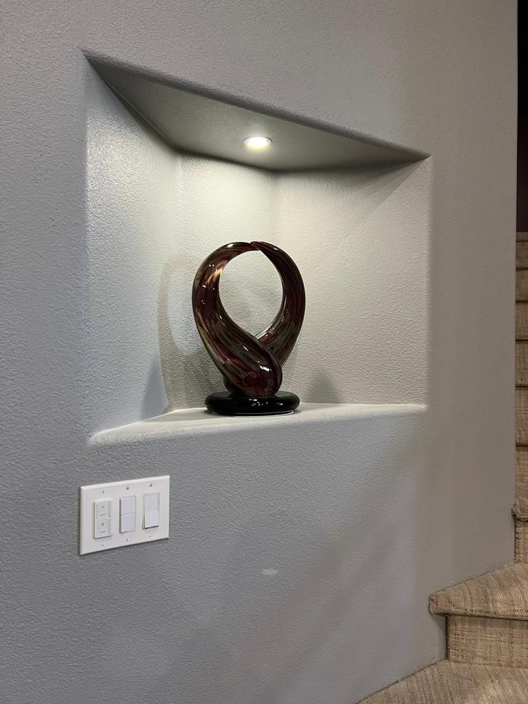 Built-in wall niche with an abstract sculpture illuminated by a spotlight. Gray textured walls and light switches below.