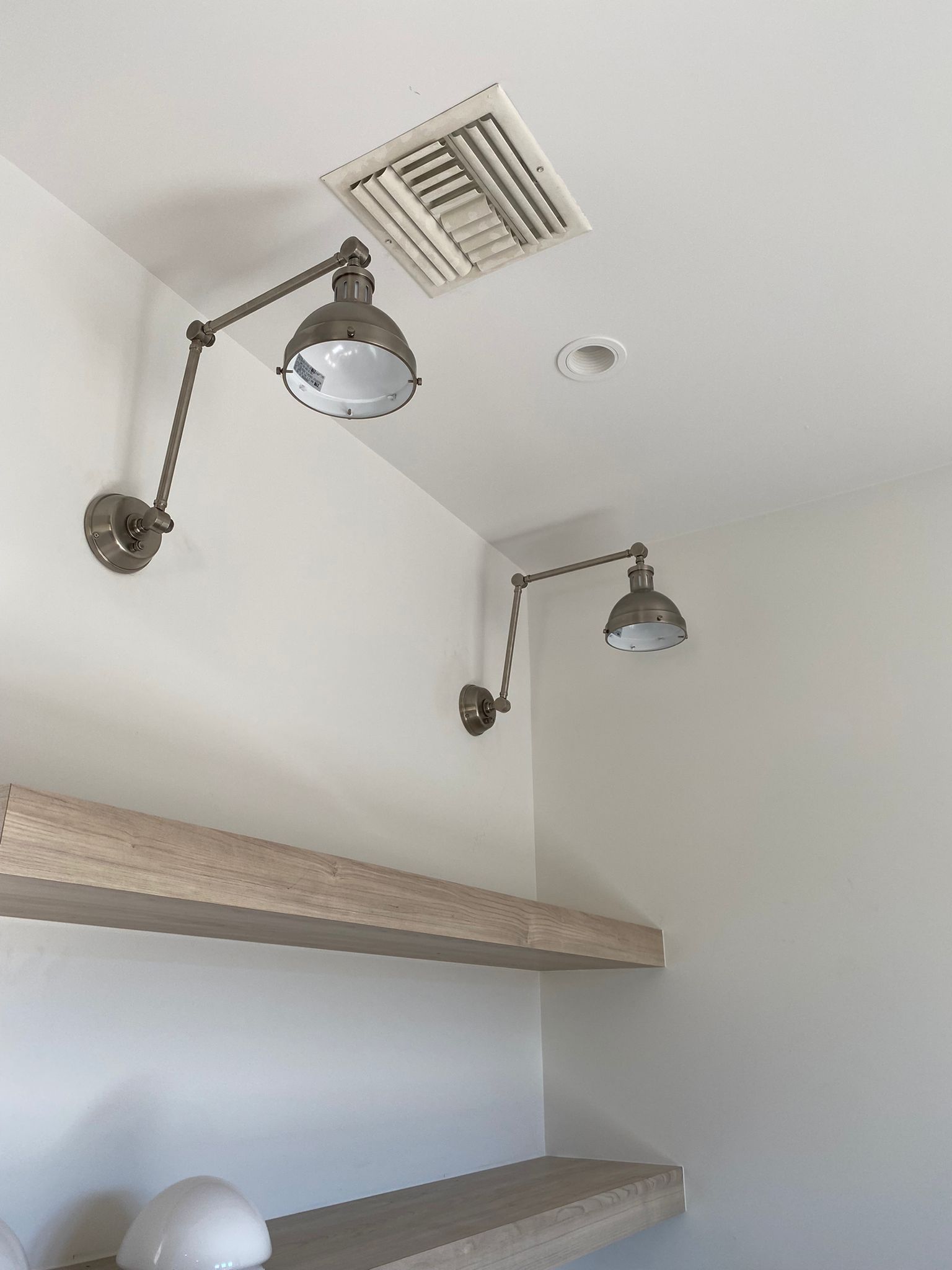 Two articulated wall lamps on a white corner, above light-colored floating shelves. A vent is in the ceiling.