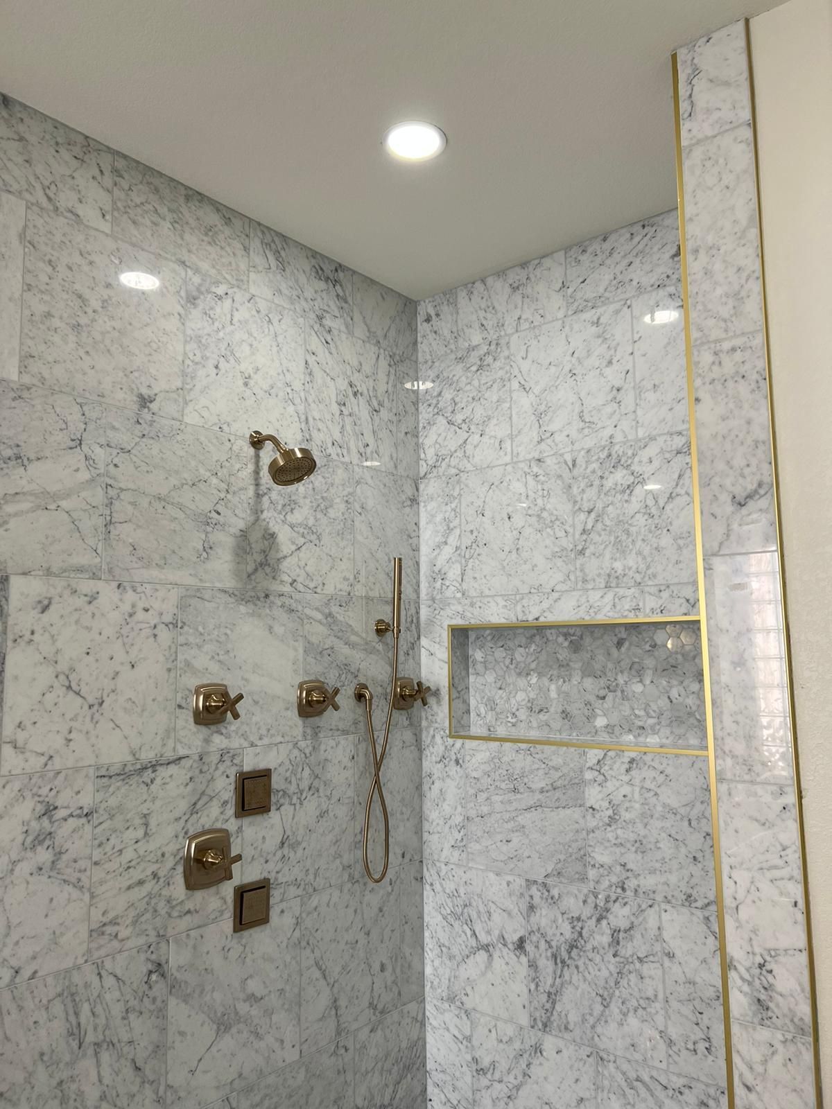 Shower with marble-like tile, gold fixtures, recessed shelf, and bright overhead light.