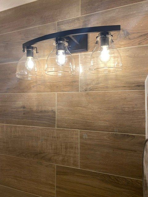 Black three-light fixture with clear glass shades mounted on wood-look tile wall.