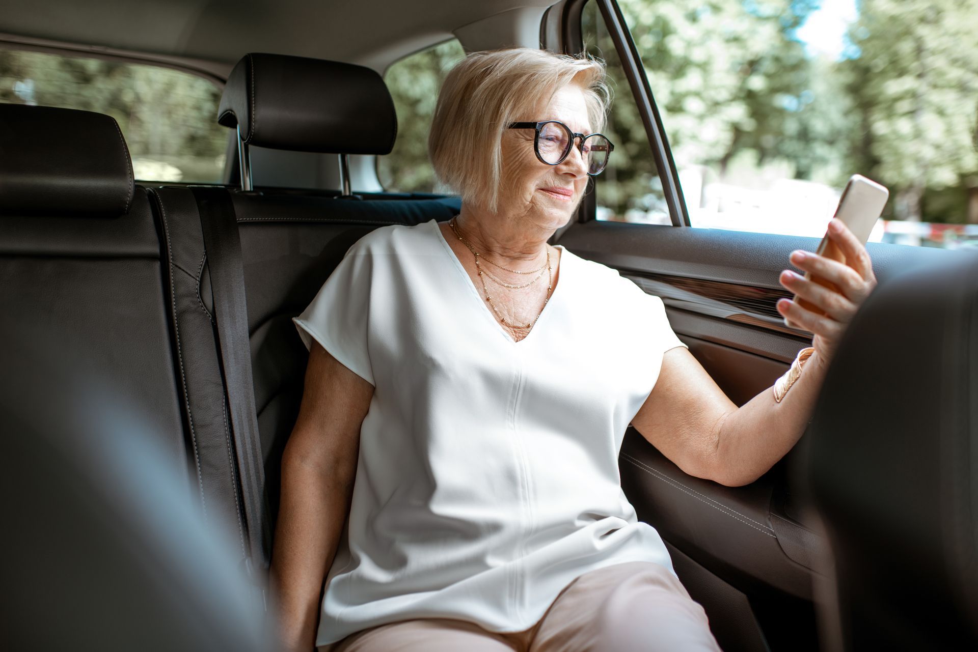 An elderly woman is sitting in the back seat of a car looking at her cell phone.