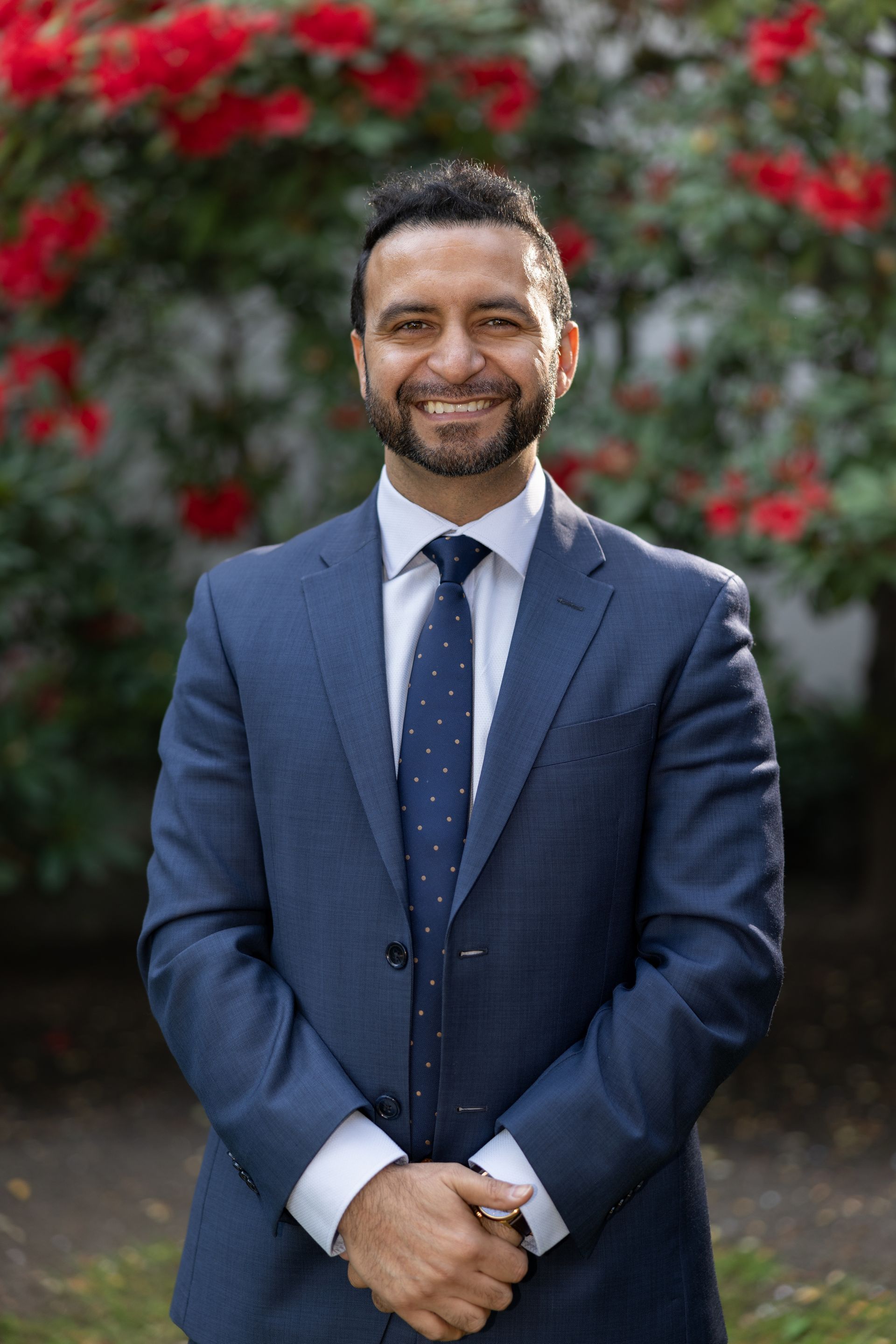 A man in a suit and tie is standing in front of a bush of red roses.