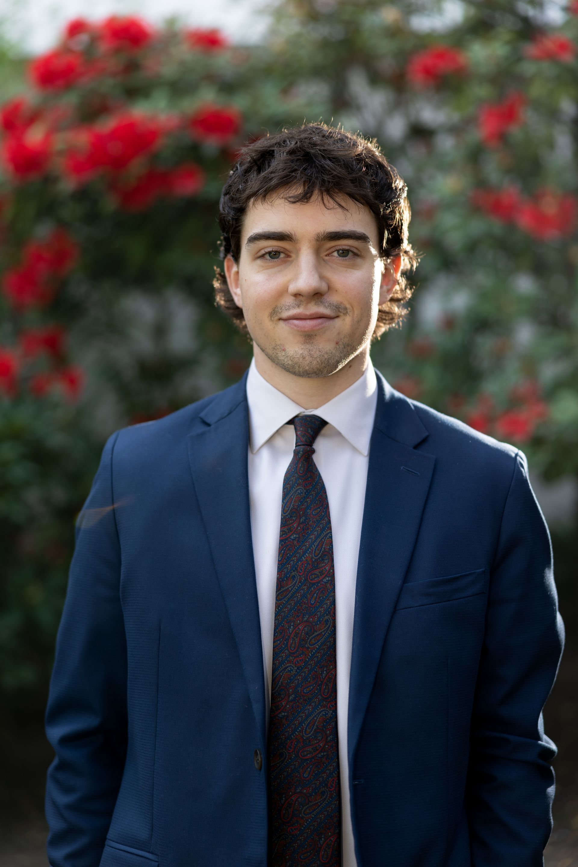 A man in a suit and tie is standing in front of a bush of red flowers.