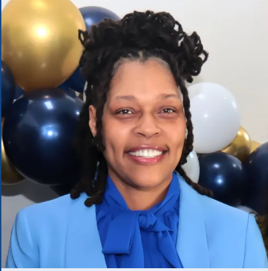Smiling person in a blue blazer and blue blouse, positioned in front of gold, blue, and white balloons.