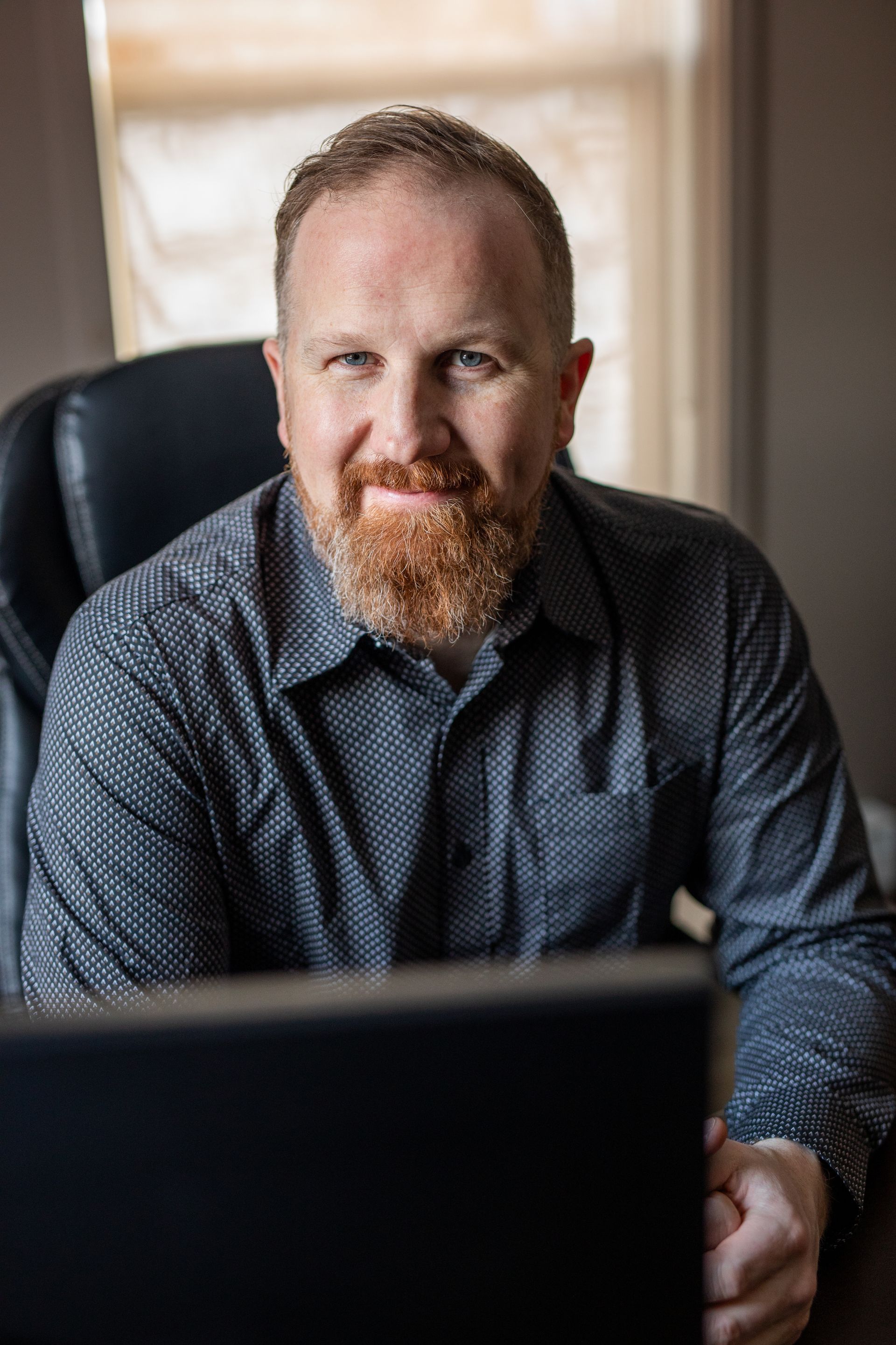 Man in a patterned shirt sitting at a desk, smiling behind a laptop in a warmly lit room