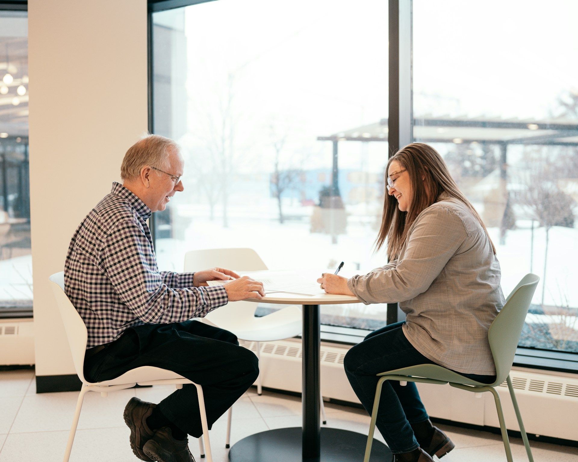 A man and woman sit at a table by a window, working. The woman writes; the man gestures with a pen.