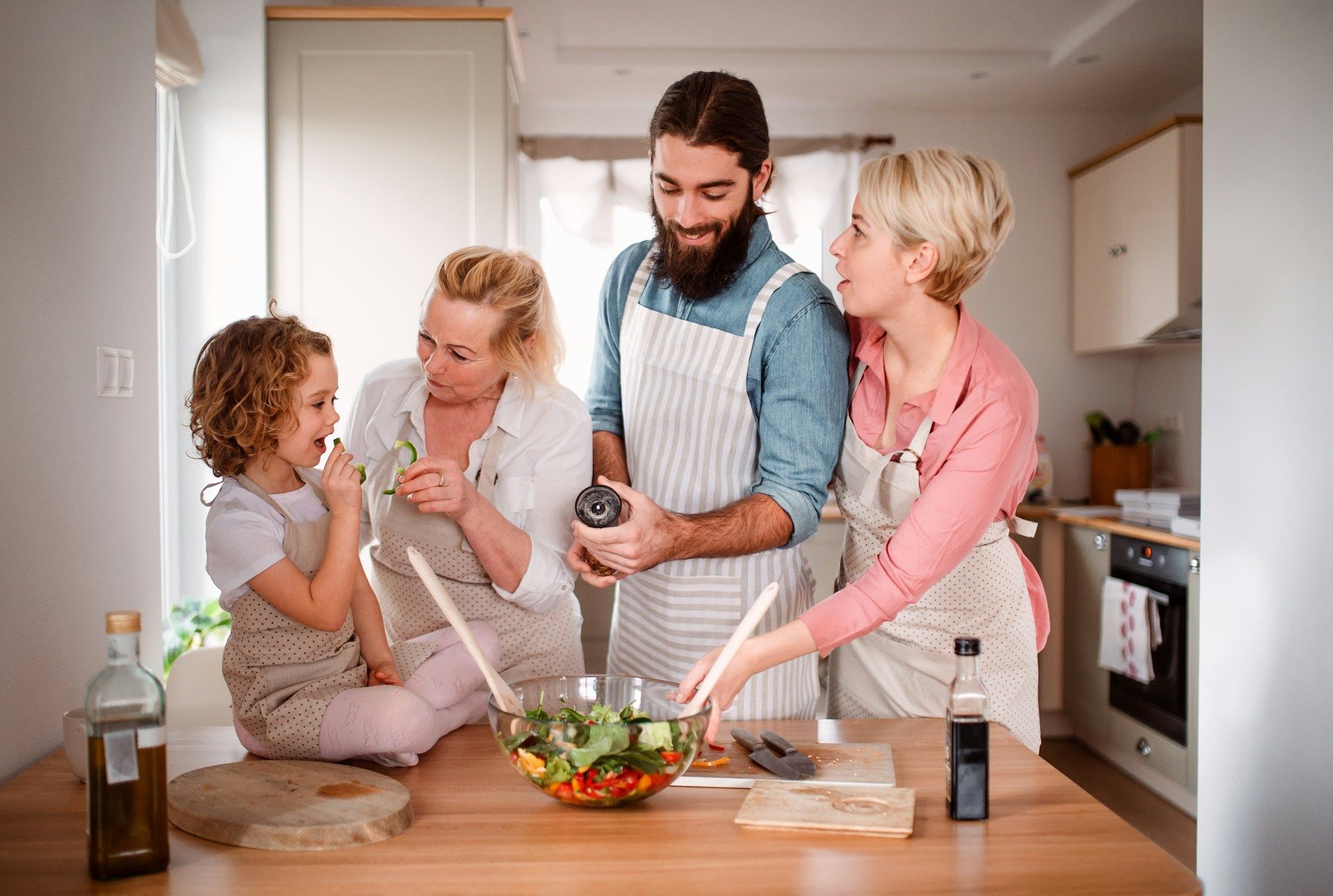 Family in kitchen preparing a salad: child, man, two women. Light-filled kitchen, wooden table, aprons.